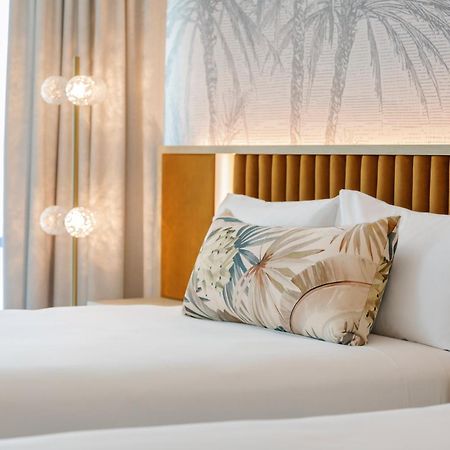 Amora Hotel Brisbane Eksteriør bilde The photo shows a stylish hotel room featuring two neatly made beds with white linens. Each bed has several pillows, one of which has a decorative cover with a tropical plant pattern. Behind the beds is a modern headboard, and the wall is decorated w