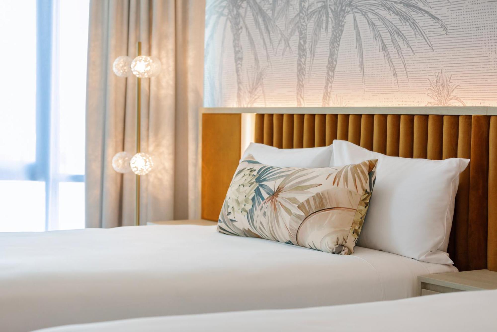 Amora Hotel Brisbane Eksteriør bilde The photo shows a stylish hotel room featuring two neatly made beds with white linens. Each bed has several pillows, one of which has a decorative cover with a tropical plant pattern. Behind the beds is a modern headboard, and the wall is decorated w