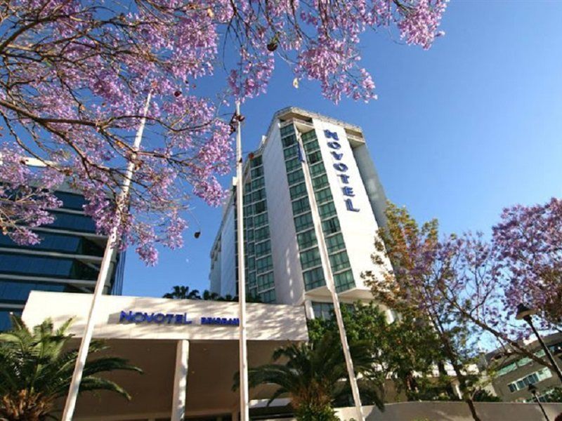 Amora Hotel Brisbane Eksteriør bilde The photo shows a modern hotel building, identified as "Novotel," rising above a landscaped area with palm trees. In the foreground, there are vibrant purple flowering trees, likely jacarandas, which add to the picturesque scene. The sky is clear and